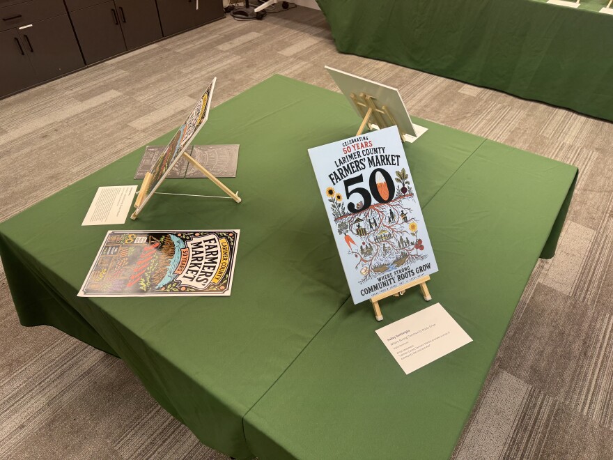 Three posters face outward in a triangle formation on a small, square table. One poster has a linocut and a rough draft next to it. Another poster is blue. The last poster is facing the wall and the front cannot be seen. In front of all three is a sheet of paper with the artist name and description.