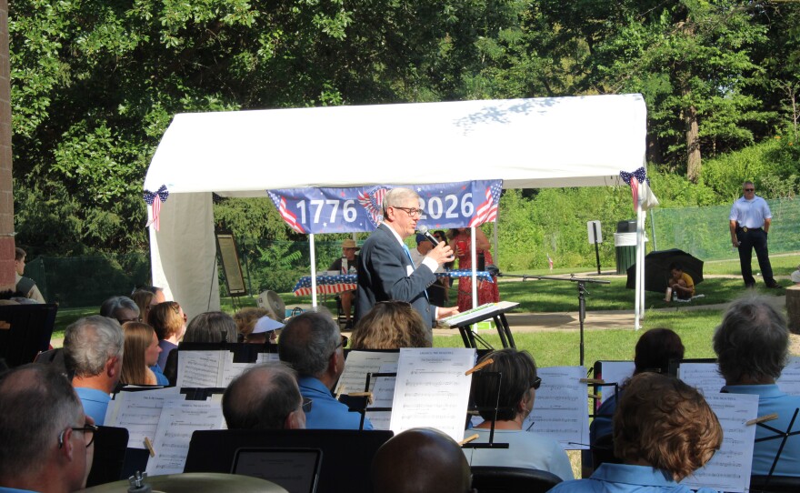 A man in a suit speaks into a microphone under a tent decorated with a "1776–2026" banner, with band members seated nearby holding sheet music.