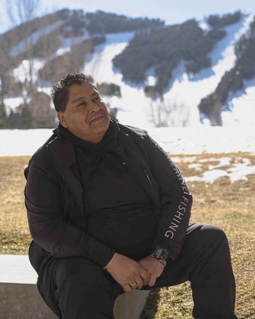 A man sits on a concrete slab in a field of dried grass. A snowy forested hillside rises above him with ski runs streaking through it.