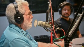 Two men sit in a radio studio. The man at left is talking and the other is listening. They are sitting at microphones.