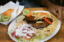 An oval white plate sits on a table. A man's hand is touching the side. The plate shows a salad with white dressing, rice and blackened fish. Beside the plate is a cardboard container with a brown, fried pastry.