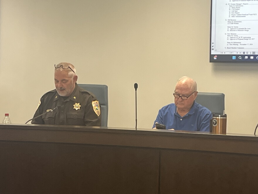 An image of a man in sheriff's department uniform and another man sitting on a dais in a meeting of the McLean County Emergency Telephone System Board in McLean County.