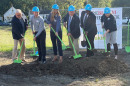 Six people holding green shovels break ground at a construction site