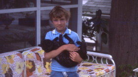 Small boy holding a black cat. Behind him is a rattan couch with floral-printed cushions.