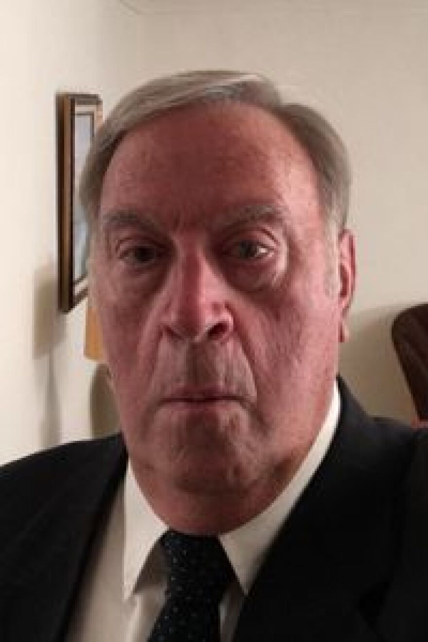 A mean wearing a suit and tie faces the camera. His lips are pursed, not quite a smile.