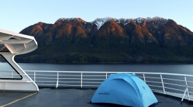 A small tent pitched on the deck of a ferry, with coastal mountains in the background.