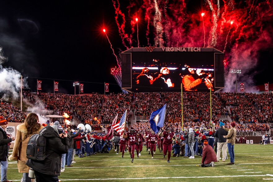 Football players in maroon jerseys run onto a field in front of a packed stadium, carrying the American and the Virginian flags. 