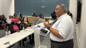 A photo of Tarrant County Commissioner Roderick Miles Jr., a Black man with short salt and pepper hair and wearing a white polo and black pants, speaks at a podium in front of a small meeting room filled with people listening to his remarks.