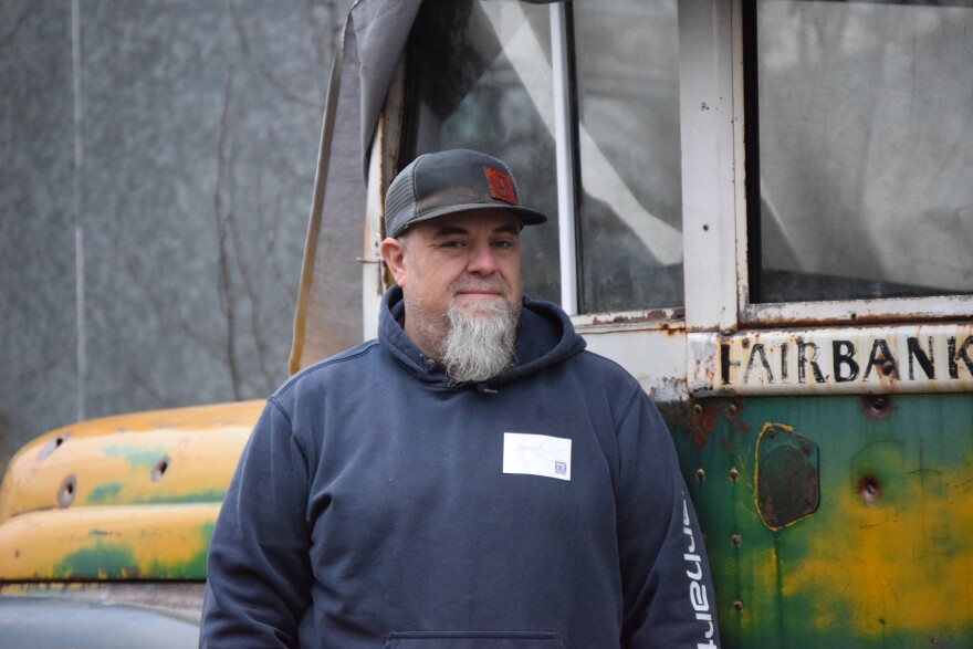 A middle-aged man with a long, gray goatee stands by the driver's side door of the green-and-yellow bus that Chris McCandless died in.