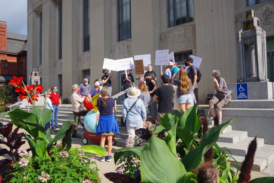  About two dozen people stand in front of Kalamazoo City Hall listening to speakers on the climate crisis and the public health implications of climate change.  There are potted flowers in the forefront of the photograph.  Signs and people in respirators and masks draw attention to the smoke from wildfires in Canada. 