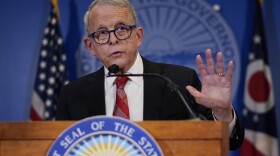 a man in blue framed glasses and a white shirt, red tie and black suit jacket talks at a podium. behind him are the american flag, ohio state flag and ohio state seal