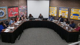 A group of people sit around a large U-shaped table in a meeting room, with colorful bulletin boards and a blank projector screen on the back wall.
