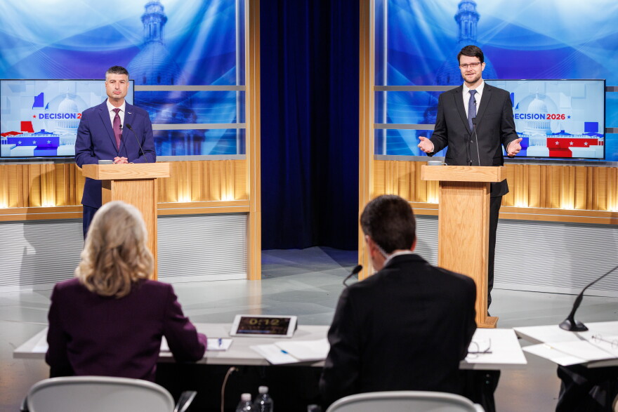 Two men in suits stand at podiums facing a man and woman sitting at a table in a TV studio
