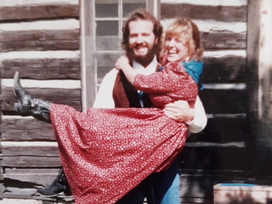 A man holds a woman in his arms. They're in front of a log cabin. She's wearing a long red dress and black cowboy boots. He's wearing a white collared white and brown vest. 