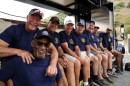 A row of men in blue T-shirts sit on a truck bed