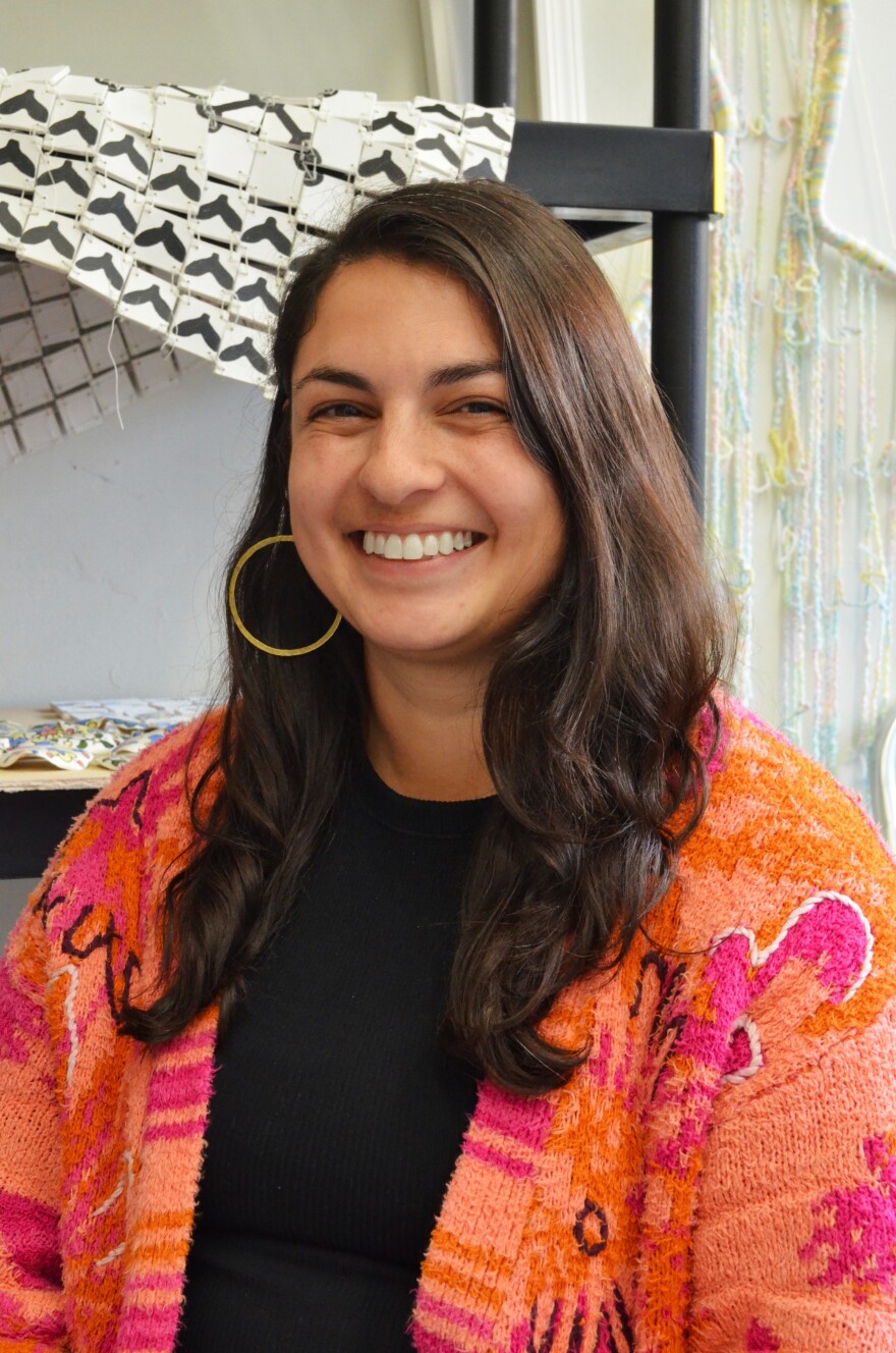 A young woman with long dark hair and a big smile, and wearing an orange and fuschia sweater, smiles at the camera. 
