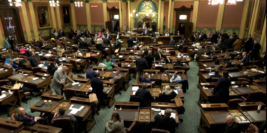 A wide-angle overhead view of the Michigan House of Representatives chamber during an active floor session. Dozens of legislators mill about, converse at desks, and move between rows of dark wood legislative desks arranged on a green carpeted floor.