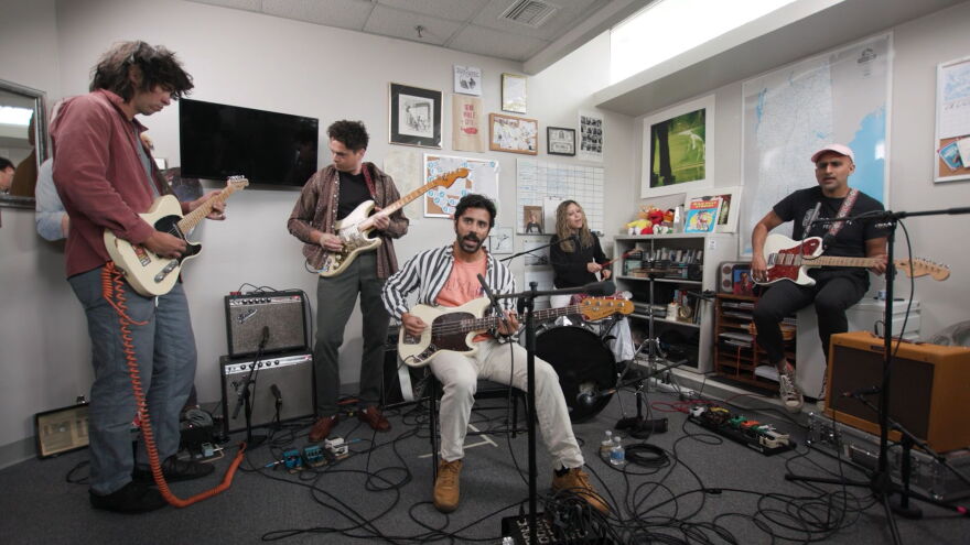 A five-piece rock band playing in a small office. Two guitarists stand on the left, a vocalists playing bass sits in the middle. A third guitarist sits on the right and a drummer plays in the back.