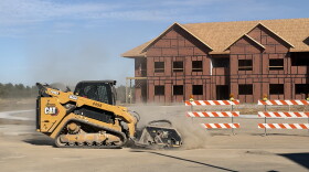 A yellow bulldozer uses a brush attachment to sand down poured concrete in front of a framed condo structure 