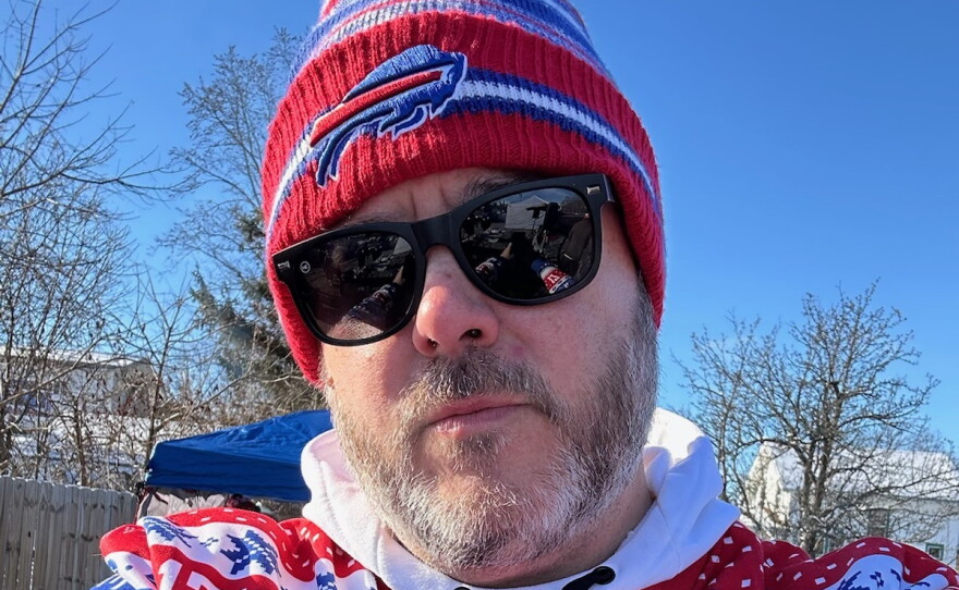 A man with a greying beard stands outside wearing dark sunglasses, a Buffalo Bills knit hat and a red, white and blue shirt.