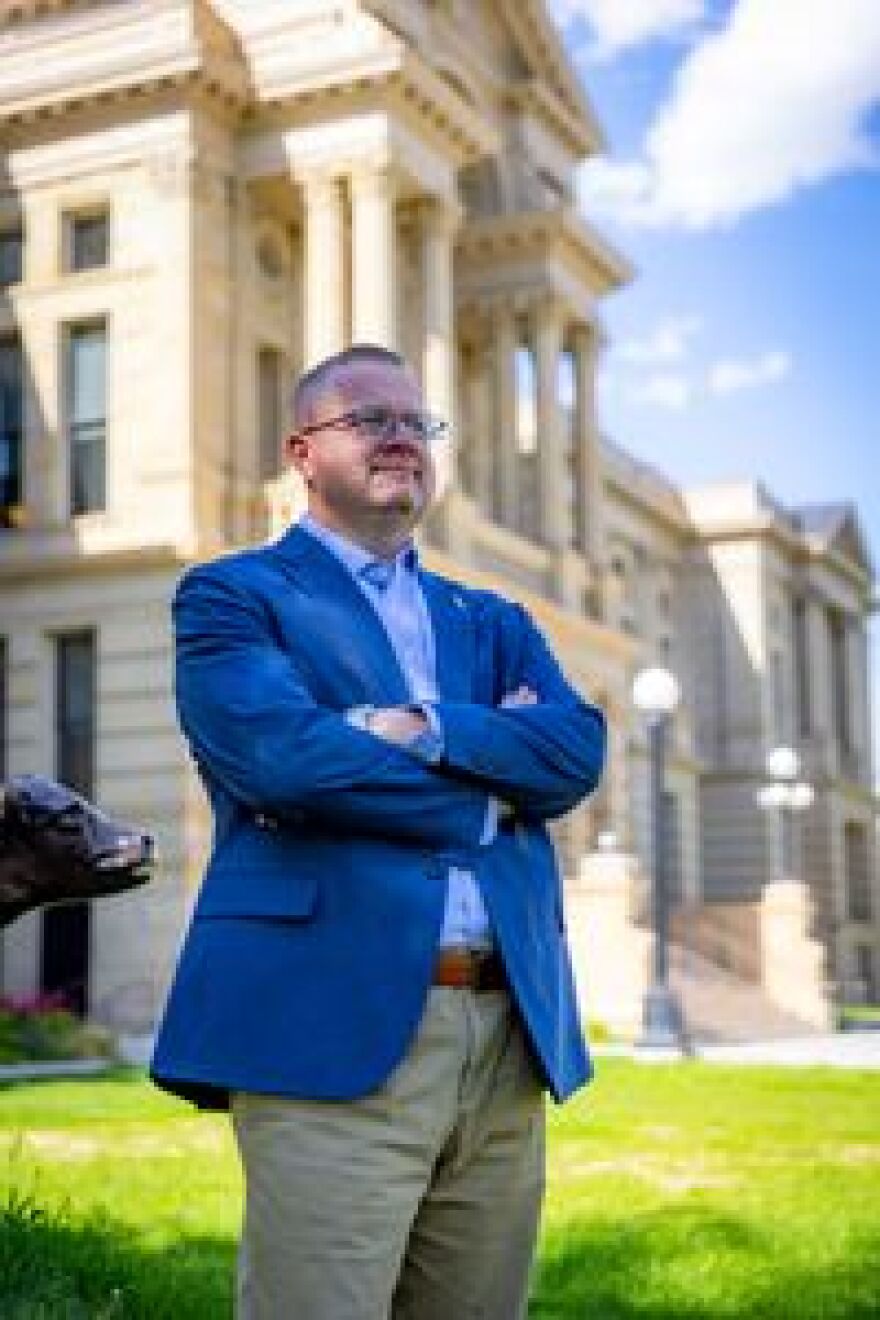 Older man in glasses and blue suit stands with arms crossed in front of capitol building