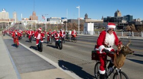 Hundreds of cyclists dressed as Santa Claus ride over a bridge with a city skyline in the background.