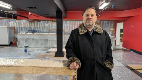 A man wearing a black coat with leopard print furry cuffs leans against a bare wood structure inside a basement that is covered in plastic sheeting