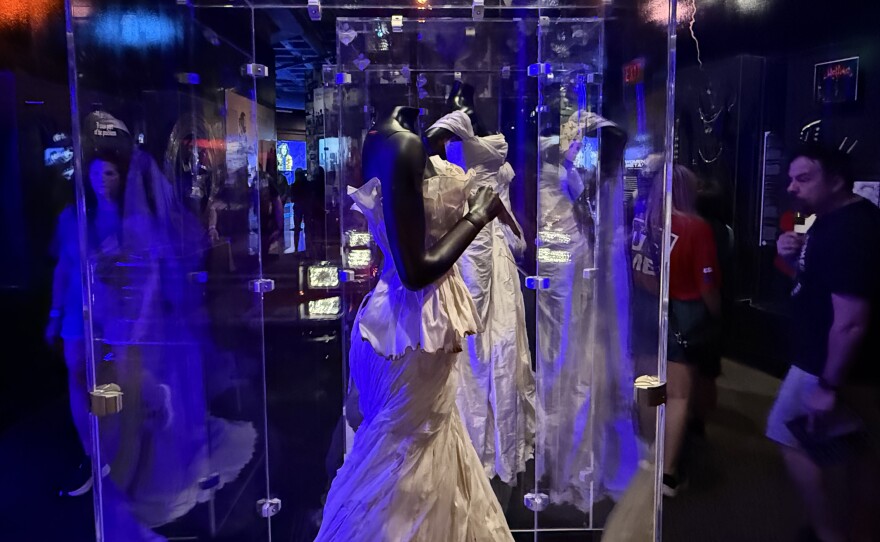 Two white gowns with singed hems are displayed in glass museum cases, while museumgoers observe in the background.
