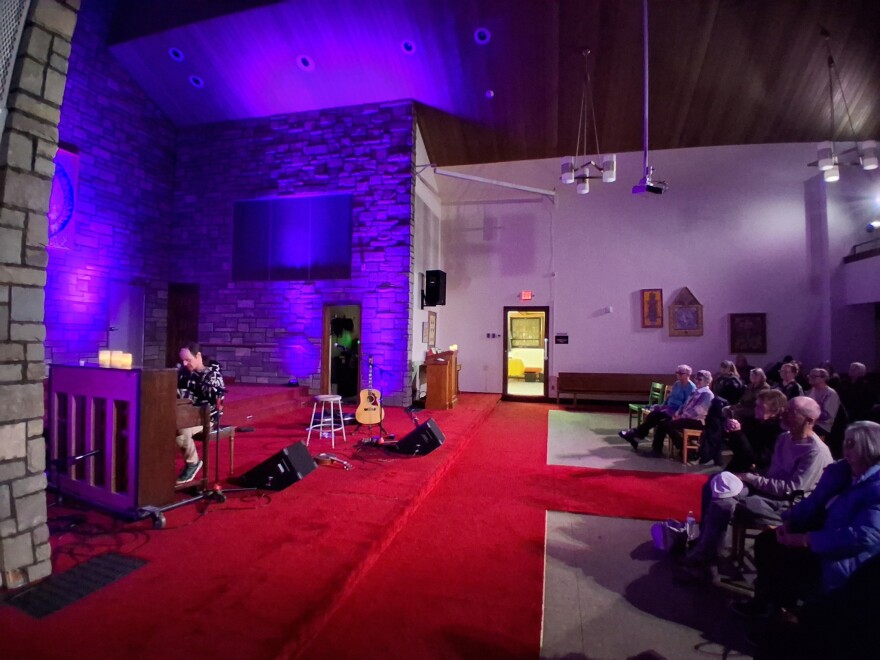 A person playing piano on a low, red-carpeted stage in front of a seated crowd