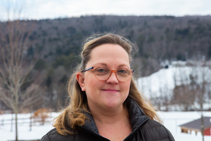 A photo of a light-skinned person wearing glasses and with blonde hair in a half-ponytail. She's wearing a jacket and standing outside with snowy mountain in the background.