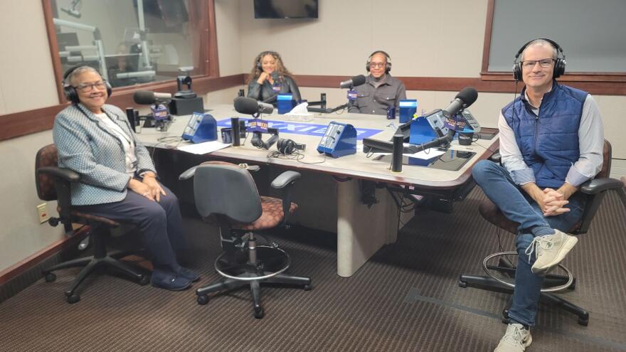 Four people wearing headphones sit at a table in a radio talk studio: a woman front left has very short grey hair and is wearing glasses, a blue and white blazer, white shirt, blue pants and blue shoes; a man front right has short dark hair and is wearing glasses, a navy puffy vest, white button-down shirt, jeans and sneakers; a woman back left has long brown hair and is wearing a black shirt; a bald man back right is wearing glasses, a grey turtleneck and a grey button-down shirt.