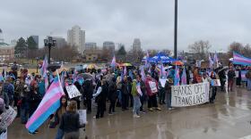 A group of people standing on the steps of the Utah Capitol, many with trans flags and signs. The largest visible sign says "hands off trans lives!"
