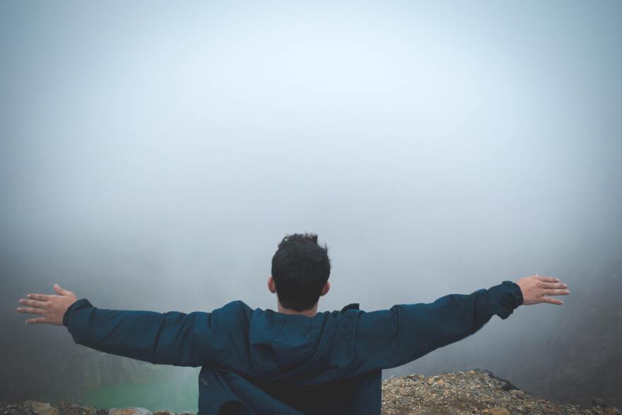 A man standing on the edge of a cliff, looking into the foggy sky.