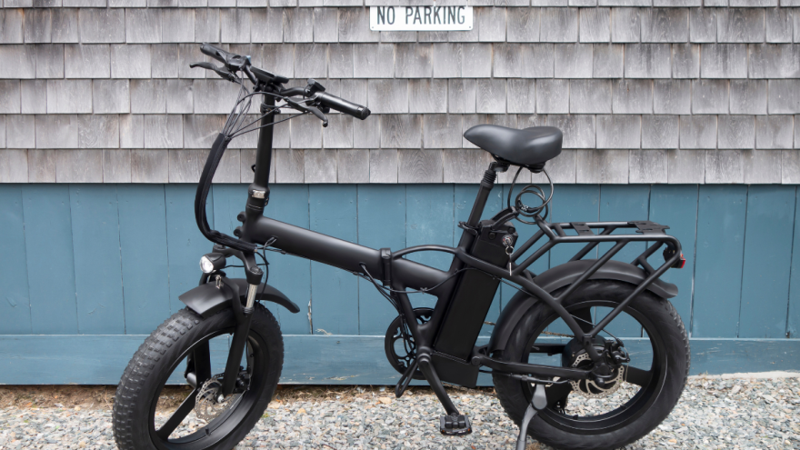 A black electric bicycle sits parked in front of a blue and gray wooden wall with a sign that says, "no parking."