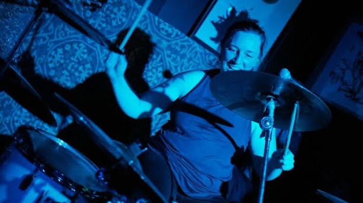 Cast in blue light, a woman smiles while playing a drum set; camera angle is at a right diagonal