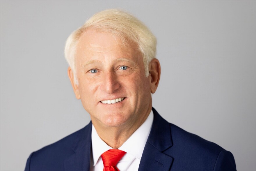 A headshot of a man with white hair, blue eyes, a blue shirt and a red tie