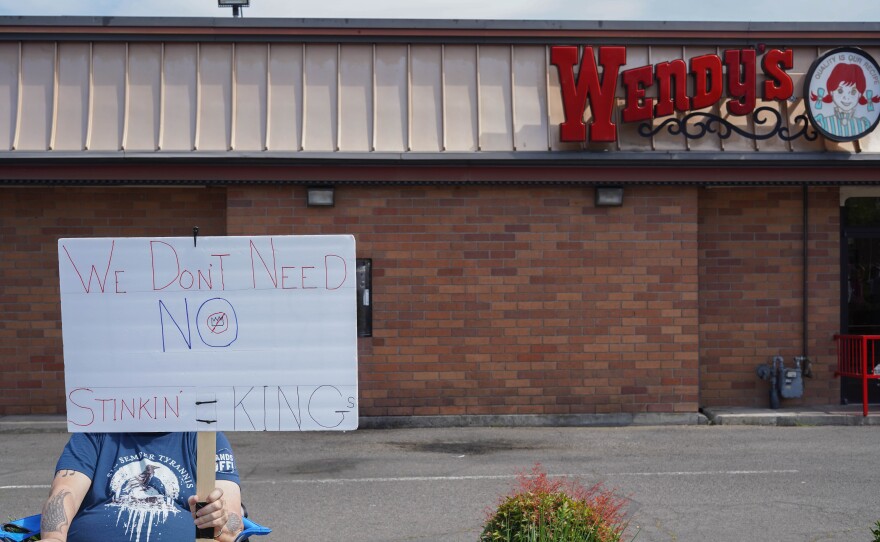 Person seated in a folding chair outside a Wendy’s holding a handwritten protest sign that reads “We don’t need no stinkin’ kings.”