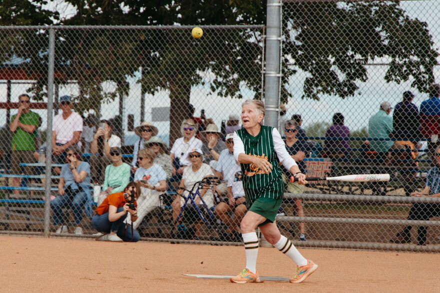 A senior woman wearing a green baseball jersey with orange text on it swings a white baseball bat at a yellow softball at home plate. She is looking up and the ball and is starting to run. Behind her is a metal fence and fans sitting on bleachers watching her.