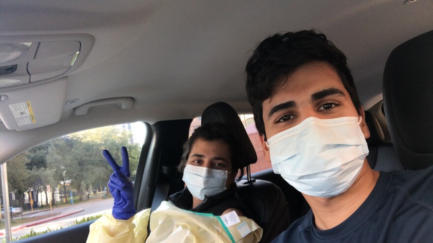 Yusuf Khan in the car and his mother Nadia Shaik. Nadia holds up a peace sign while wearing a medical gown, gloves and face mask.