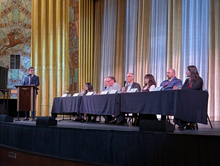 Seven people sit behind black tables on a stage, a man in a suit on the left stands behind a podium