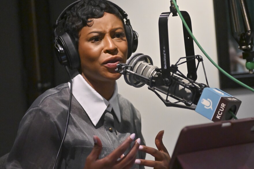 A woman sits inside a radio studio at a microphone. She is talking and gesturing impassionedly with both hands.