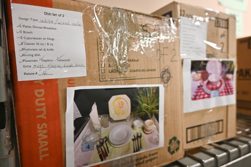 Two cardboard boxes are seen on a wire rack shelf. They are labeled "Dish set of 2." There are pictures taped to them showing a place set with silverware that describes the contents of the box.