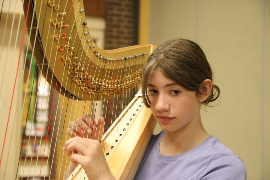 A young girl with brown hair in a ponytail and a violet-colored shirt has her hands on the strings of a golden harp. She's looking down at her music, which is not shown in the photo. 