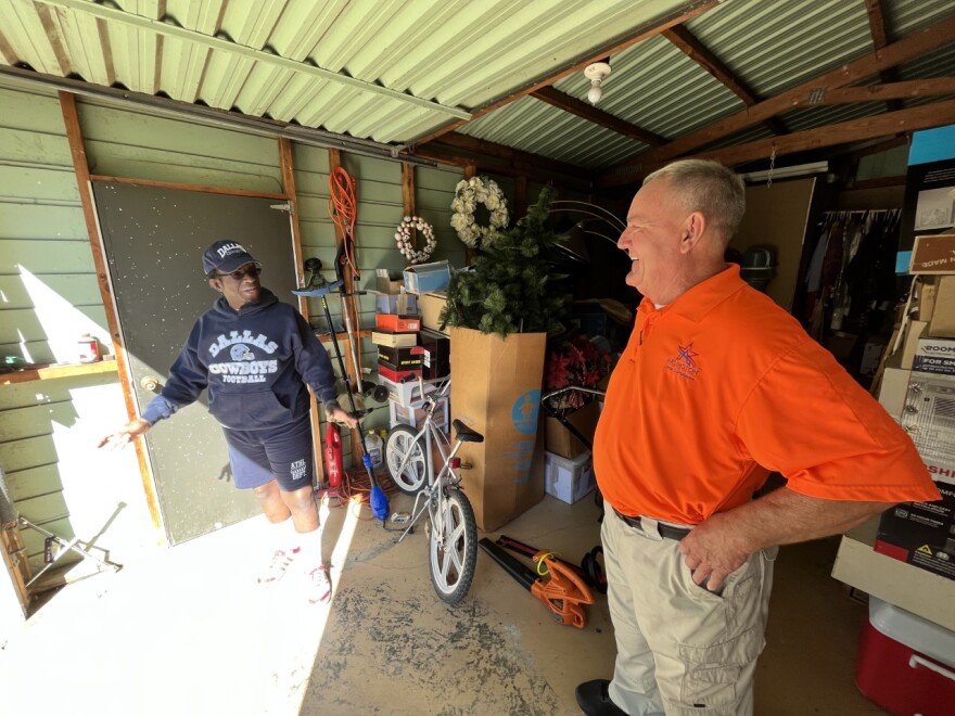 A man wearing a dark blue Dallas Cowboys sweatshirt, cap and knee-length shorts with knee-high socks talks to a man with a neon orange shirt and khakis. They're standing inside a green-painted shed that contains a bicycle, assorted boxes and an orange yard tool. The man in the Cowboys merchandise, Chris Lane, has his arms slightly spread out as he speaks, while the man in neon, Ron McCall, smiles at him with his hands on his hips and partially tucked into his pant pockets.