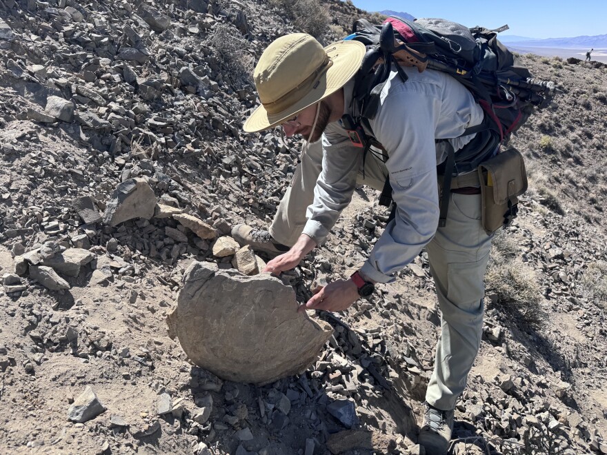A man in a hat and long sleeved shirt bends over to look at a large, round rock.