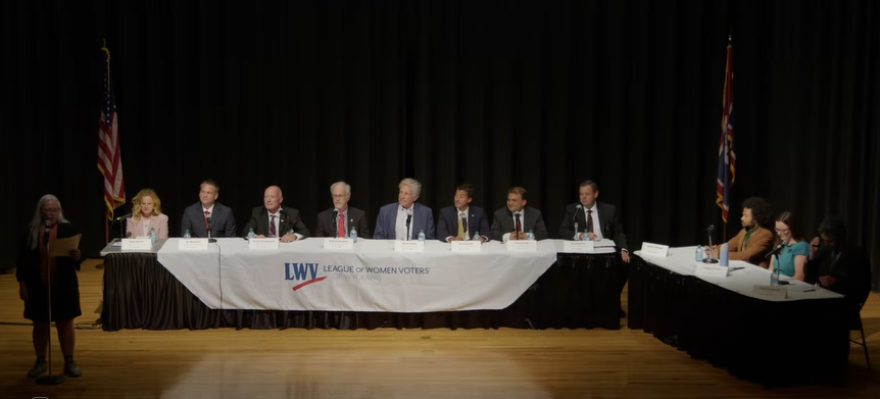 Seven men and a woman sit at along table. They're flanked on the stage by a table of a few journalists and a woman standing while speaking into a microphone. The ensemble looks a bit like "The Last Supper."