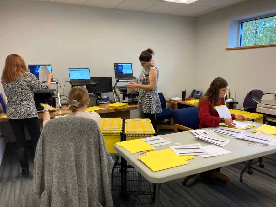 Four people work in a room with computers along the back wall and tables covered in papers. 