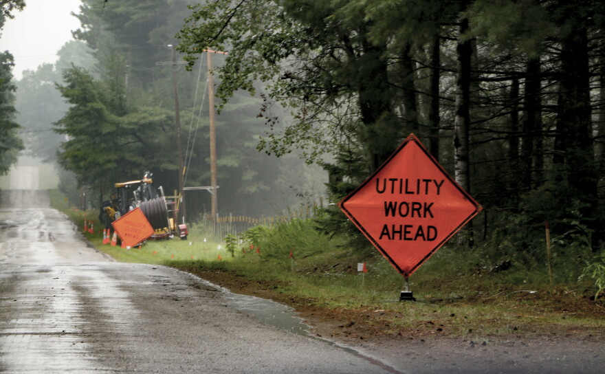 A stock photo shows an orange roadside sign warning of utility work ahead, with a heavy machine in the background carrying a large spool of cable.