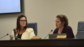 Two women sit at a desk with microphones. 