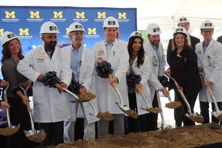 A group of people are lined up wearing white lab coats and hard hats with University of Michigan branding. They are all smiling while holding shovels, digging up a mound of dirt to symbolically break ground for the construction project.